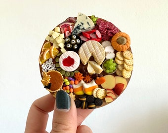 Op de afbeelding: Een miniatuur Halloween-kaasplank met een verscheidenheid aan kazen, crackers, fruit en snoepjes. De plank is gemaakt van hout en is versierd met herfstaccenten.