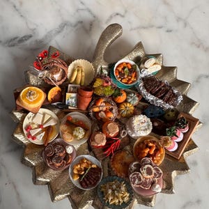 May include: A gold-colored, leaf-shaped serving tray filled with miniature food items. The tray holds various dishes, including pasta, steak, desserts, and fruit. The food items are arranged on small plates and bowls, creating a visually appealing display.
