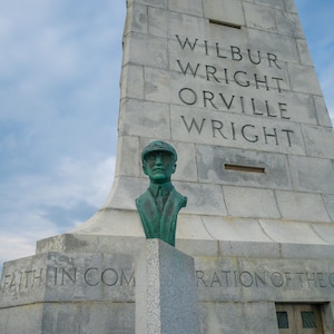 Puede incluir: Un busto de bronce de Wilbur Wright, uno de los hermanos Wright, se encuentra frente a un monumento de piedra. El monumento está inscrito con los nombres "Wilbur Wright, Orville Wright" y la frase "Fe en común, Inspiración de los conquistados".