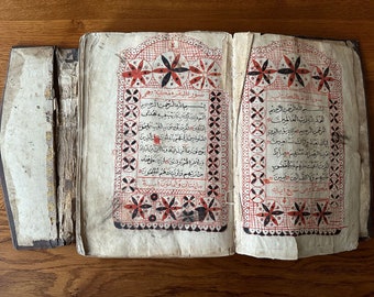 May include: An open book with Arabic text and intricate red and black geometric designs on the pages. The book is old and worn, with some pages missing.