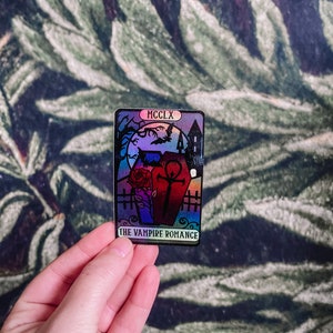May include: A holographic sticker with a black and white illustration of a coffin, a rose, and a cross. The sticker has the text "MCCCLX" and "THE VAMPIRE ROMANCE" on it.