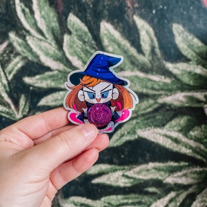 May include: A sparkly witch sticker with a blue hat and a pink and purple magic orb. The sticker is on a green leafy background.