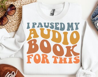 May include: White sweatshirt with the text "I paused my audiobook for this" in a retro font with blue, orange, and yellow colors.