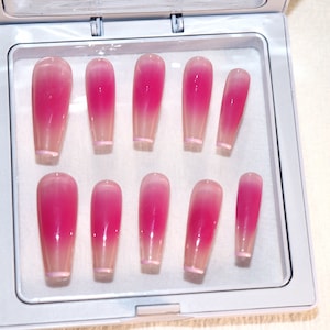 Puede incluir: Un juego de diez uñas postizas de color rosa y blanco degradado en un contenedor de plástico transparente.