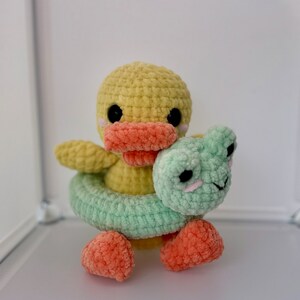 Peut inclure: Un canard jaune en crochet tenant une bouée en crochet verte et blanche. Le canard a des yeux noirs et un bec orange.