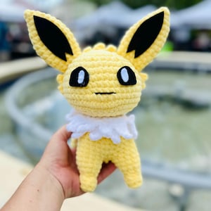 May include: A handmade crochet toy of a yellow creature with large ears, black ear tips, and black eyes. It has a white ruffled collar. The toy is held in a hand. The character is from a popular animated series.
