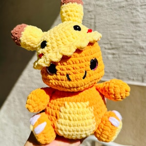 May include: A crocheted plush toy in the shape of a yellow and orange Pokemon character. The toy has large eyes and a small mouth. It is wearing a yellow crocheted hat.