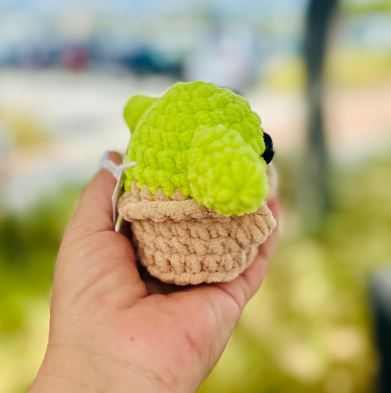 May include: A small green crocheted plush toy in the shape of a character from the Star Wars franchise. The toy is sitting in a brown crocheted pot.