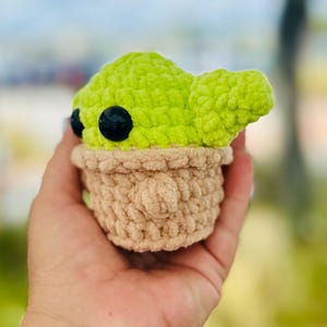 May include: A crocheted plush toy of a green character with large black eyes. The character is wearing a brown crocheted hat and has a small, pointed ear.