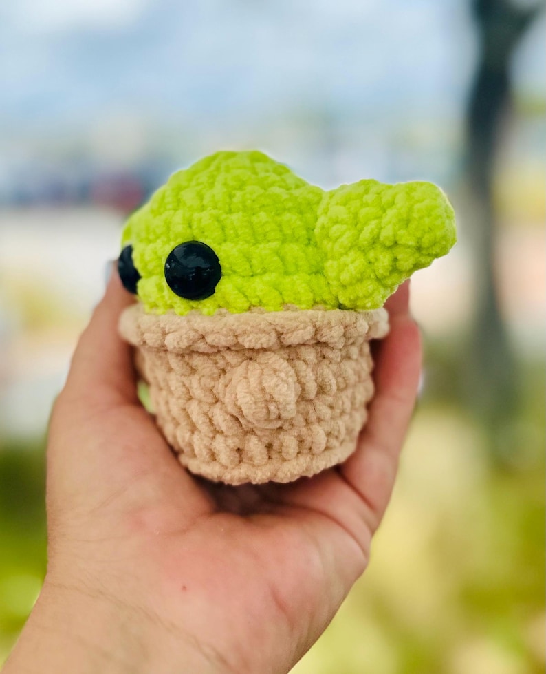 May include: A crocheted plush toy of a green character with large black eyes. The character is wearing a brown crocheted hat and has a small, pointed ear.