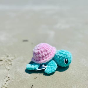 Könnte beinhalten: Ein rosa und blaues gehäkeltes Stofftier in Form einer Schildkröte auf einem Sandstrand.