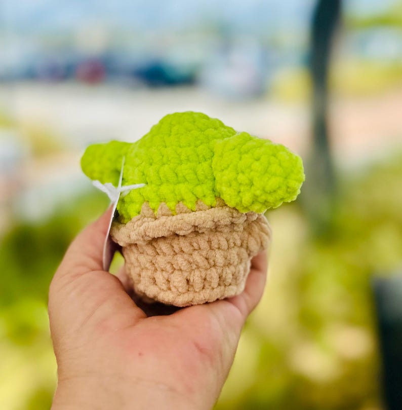 May include: A crocheted plush toy shaped like a character from the Star Wars franchise. The toy is green and brown and has a pot-like base.