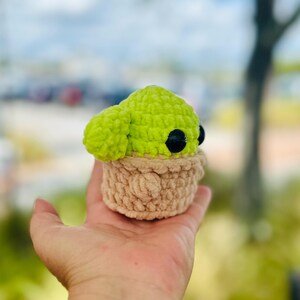 May include: A crocheted green and brown plush toy resembling a character from the Star Wars franchise. The toy has large black eyes and is sitting in a person's hand.
