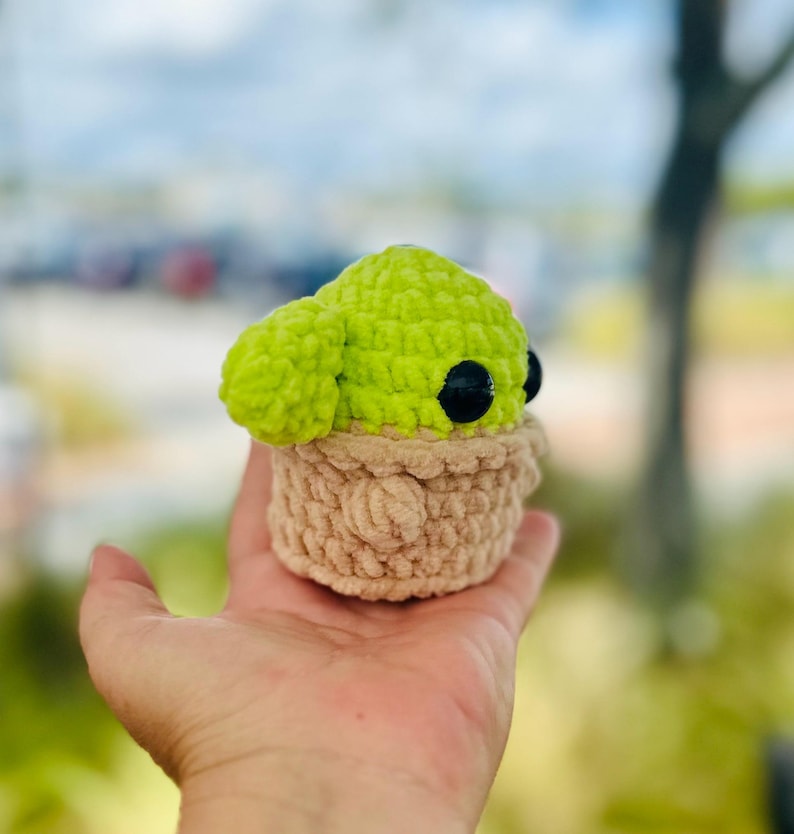 May include: A crocheted green and brown plush toy resembling a character from the Star Wars franchise. The toy has large black eyes and is sitting in a person's hand.