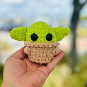 May include: A small green and brown crocheted plush toy resembling a character from the Star Wars franchise. The toy has large black eyes and a small mouth.