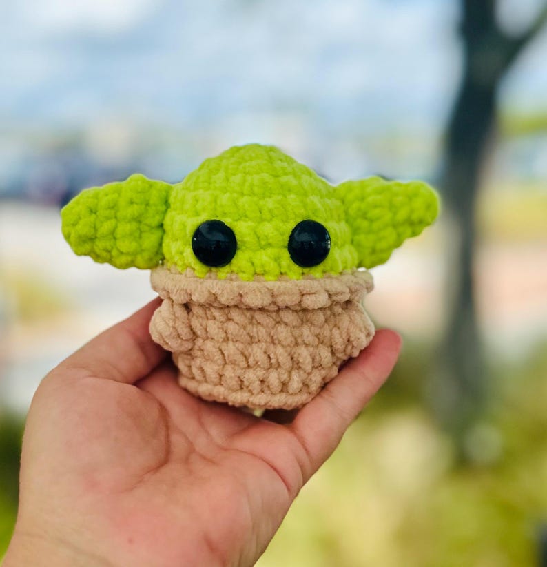 May include: A small green and brown crocheted plush toy resembling a character from the Star Wars franchise. The toy has large black eyes and a small mouth.