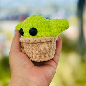 May include: A crocheted plush toy of a green character with large black eyes, wearing a brown crocheted hat. The toy is being held in a hand.