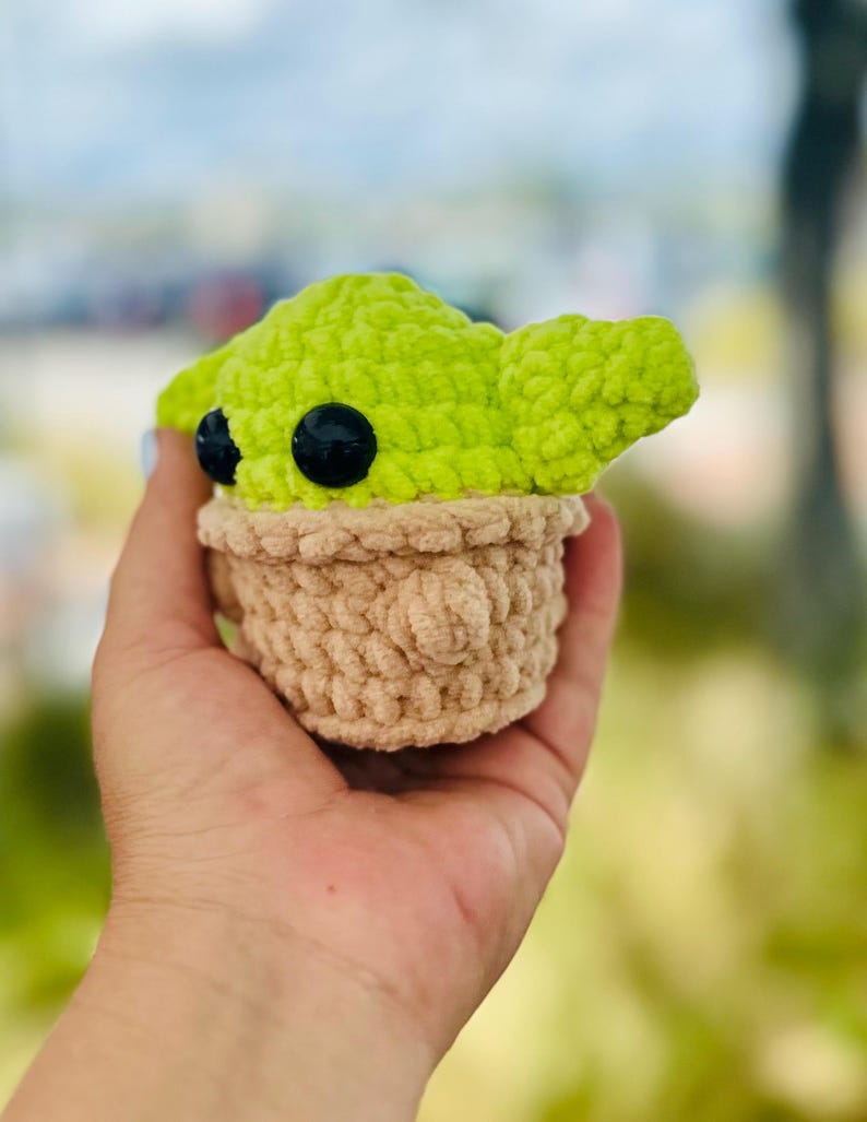 May include: A crocheted plush toy of a green character with large black eyes, wearing a brown crocheted hat. The toy is being held in a hand.