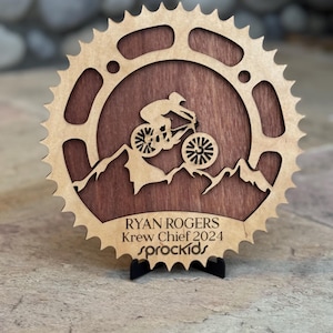 May include: A wooden mountain bike award with a brown wood background and a silhouette of a mountain biker. The award is shaped like a bicycle chainring and has the text "RYAN ROGERS Krew Chief 2024 sprockets" on the bottom.