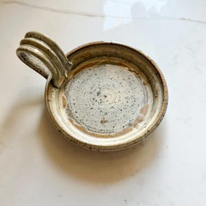 May include: A round, shallow ceramic dish with a speckled off-white glaze and a brown rim. The dish features a single, arched handle on one side, constructed with three distinct, stacked ridges that provide a textured grip for the user.