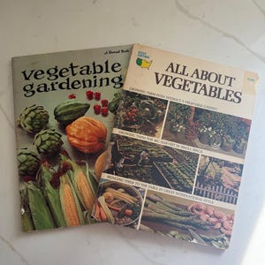 Puede incluir: Se muestran dos libros de jardinería antiguos titulados "vegetable gardening" y "All About Vegetables". El primer libro muestra ilustraciones de verduras como alcachofas, maíz y tomates. El segundo libro tiene imágenes de jardines y verduras.