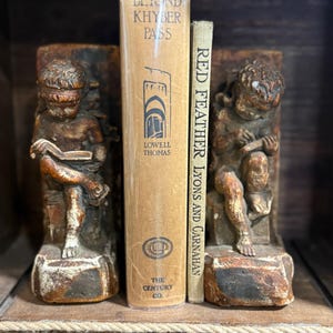 Könnte beinhalten: Antike Buchstützen mit geschnitzten Cherubinen, die zwei alte Bücher flankieren. Die Bücher tragen die Titel "Beyond Khyber Pass" und "Red Feather Lyons and Carnahan". Die Buchstützen und Bücher stehen auf einem Holzregal.