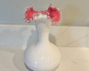 May include: A white glass vase with a bulbous base and a slender neck. The top of the vase features a ruffled, petal-like design in pink with a clear, ruffled edge. The vase is likely for decorative purposes.