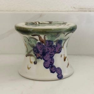 May include: A ceramic wine bottle stopper with a white base and a flared shape. The stopper is decorated with a cluster of purple grapes and green leaves. The top rim is green.