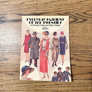 May include: A book cover titled "Everyday Fashions of the Twenties" featuring illustrations of people in 1920s clothing. The cover includes text "As Pictured in Sears and Other Catalogs" and "Edited by Stella Blum". The cover is on a wooden surface.