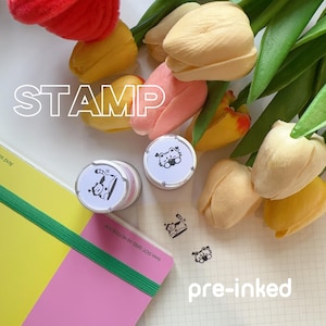 May include: A set of four pre-inked stamps featuring a cartoon cat design. The stamps are packaged in clear plastic containers and are displayed on a white gridded surface with a pink and yellow notebook. The image also includes the text "STAMP" and "pre-inked".