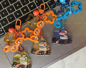 May include: Keychains featuring race car driver designs. The keychains have orange, yellow, and blue star and chain accents. Each keychain has a small race car driver graphic with text. The keychains are on a laptop keyboard.