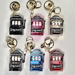 May include: Set of ten colorful keychains shaped like slot machines with the text "Smoonypaws All we need is cat" and "Lucky Jackpot".