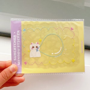 May include: A yellow and pink suncatcher window sticker featuring a white cat holding a blue balloon. The sticker has a scalloped border and the text "SUNCATCHER * WINDOW STICKER" is printed on the left side. The text "#SMOOTHYPAWS" is printed on the right side.