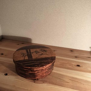 May include: A round wooden box with a dark brown finish and a natural wood grain texture. The lid features a decorative dark brown illustration of bamboo stalks and leaves set against a lighter brown, textured background surface.