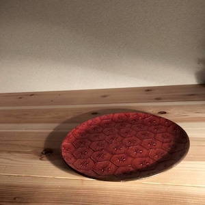 May include: A round, deep red plate featuring a repeating embossed hexagonal pattern across its surface. Each hexagon contains a small, centered star-like floral motif. The plate has a smooth, slightly glossy finish and a raised outer rim.
