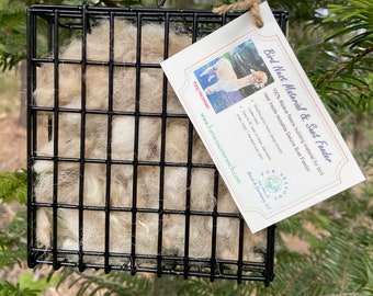 May include: A black metal bird feeder filled with light brown alpaca fleece, hanging from a chain. A tag reads "Bird Nest Material & Suet Feeder" with text and an image of an alpaca. The feeder is surrounded by green pine needles.