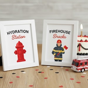 May include: Two framed prints with white frames. One says "Hydration Station" with a red fire hydrant graphic. The other says "Firehouse Snacks" with a firefighter graphic. A toy fire truck and cake are also visible.
