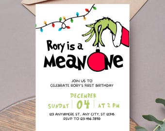 May include: A white invitation card with the text "Rory is a Mean One" in black. The design includes a green hand holding a red ornament, and a string of colorful lights. The invitation is for a first birthday party on December 4th at 2 PM.