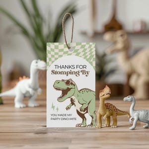 May include: A gift tag with a green and white checkered pattern and the text "Thanks for Stomping By". It features a cartoon dinosaur wearing sunglasses. The tag is attached to a brown string. Toy dinosaurs are in the background.