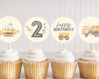 May include: Cupcakes with toppers featuring construction vehicles and the number 2. The toppers have a light yellow background with a bulldozer, a number 2, a dump truck, and the words "happy birthday".