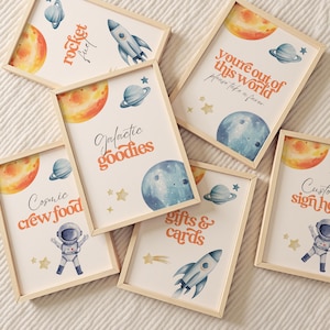 Może przedstawiać: Kolekcja oprawionych wydruków o tematyce kosmicznej. Każdy wydruk zawiera akwarelowe ilustracje planet, rakiet, astronautów i gwiazd. Tekst zawiera zwroty takie jak "Rocket Fuel" i "Galactic Goodies". Ramki wykonane są z jasnego drewna.