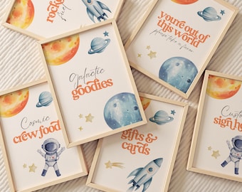 May include: A collection of framed space-themed prints. Each print features watercolor illustrations of planets, rockets, astronauts, and stars. Text includes phrases like "Rocket Fuel," "Galactic Goodies," and "Cosmic Crew Food." The frames are made of light-colored wood.
