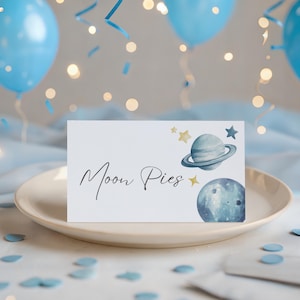 May include: A white card with the words "Moon Pies" in elegant script, decorated with watercolor planets and stars. The card sits on a beige plate, with blue confetti and balloons in the background.