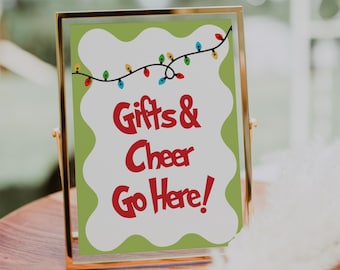 Könnte beinhalten: Ein goldgerahmtes Schild mit dem Text "Gifts & Cheer Go Here!" in Rot auf weiß-grünem Hintergrund. Eine Girlande aus bunten Weihnachtslichtern verziert die Oberseite.
