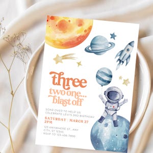 Peut inclure: Une invitation sur le thème de l'espace, de style aquarelle. Elle présente une grande planète orange, une planète bleue avec des anneaux, une fusée et un astronaute tenant un ballon. Le texte dit "Three two one... blast off".