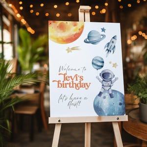 Könnte beinhalten: Eine Holzstaffelei zeigt ein farbenfrohes Geburtstagsschild zum Thema Weltraum. Das Schild zeigt eine Aquarellillustration von Planeten, Sternen, einer Rakete und einem Astronauten, der einen Ballon hält. Der Text lautet: "Willkommen zu Levis Geburtstag, lasst uns feiern!"