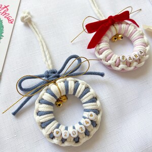 May include: Two macrame Christmas wreath ornaments. One is white and blue with gold bells and the year 2024 spelled out in beads. The other is pink and white with the name "SARA" spelled out in beads and a red bow.
