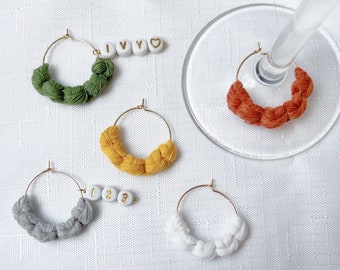 May include: Five different colored wine glass charms with gold hoops and white beads. The beads have letters and numbers on them. The charms are on a white background.