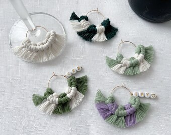 May include: Macrame wine glass charms in various colors, including white, green, and purple. Each charm features a gold hoop and a decorative tassel design. Some charms have beaded name tags with numbers or names.