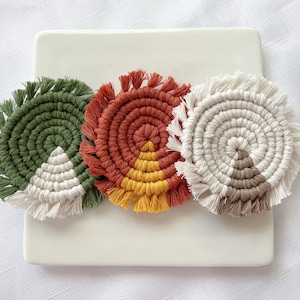 May include: Three round crocheted coasters with fringe. The coasters are green, orange, and white with a brown center. The coasters are arranged in a circle on a white surface.